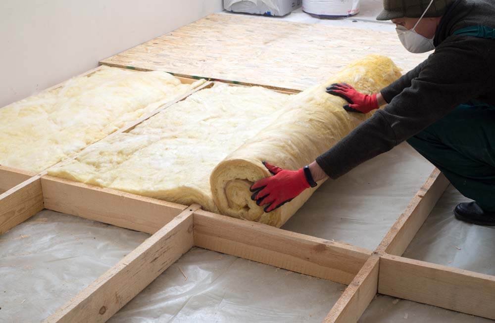 Ein Handwerker rollt eine Dämmmaterialrolle auf dem Boden aus, um den Kellerboden vor Wärme zu dämmen.